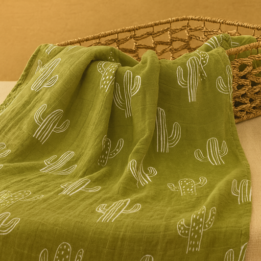 Olive green boho muslin baby swaddle with white cactus print draped over a woven basket in a warm neutral setting.